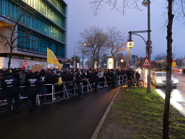 Eine große Gruppe von Menschen protestiert vor einem Gebäude in Berlin und hält Schilder mit Barrikaden, Fahrrädern, Laternen, Schildern, Bäumen und Gras im Vordergrund.