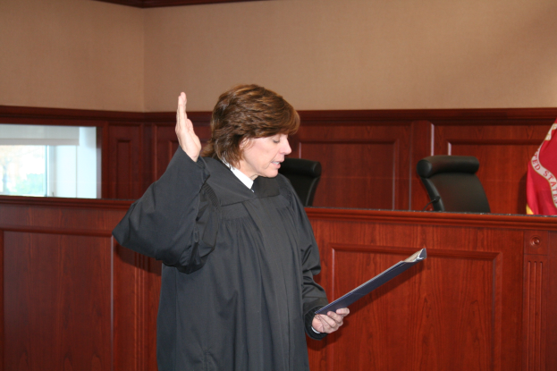 Eine Frau in einer schwarzen Robe steht an einem Richtertisch und hält eine Akte in der Hand, mit einem Mikrofon und einer Flagge auf dem Tisch, Stühle dahinter und eine Fensterwand im Hintergrund.