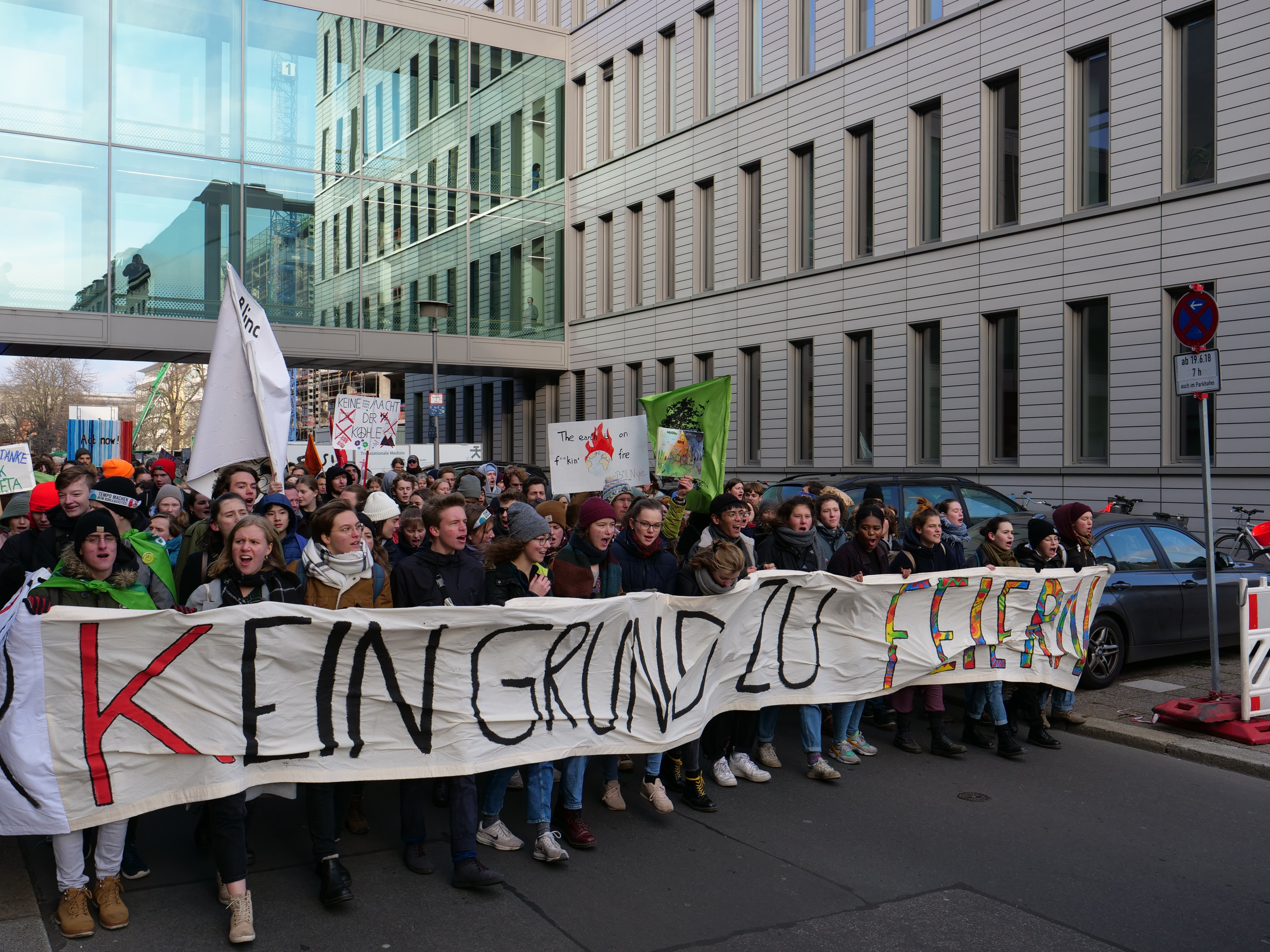 Eine Gruppe von Menschen steht auf einer Straße und hält ein Transparent mit der Aufschrift "Kein Grind Lo Feuer" hoch, mit Gebäuden, Bäumen, Polen, Schildern, Fahrzeugen und einem klaren blauen Himmel im Hintergrund.