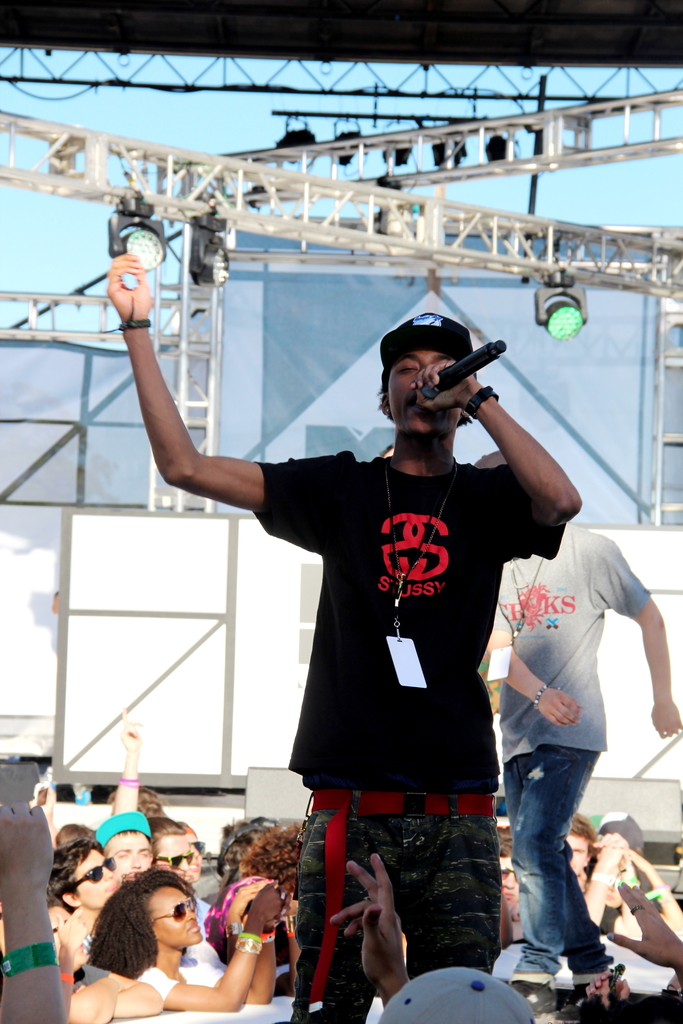 Eine Person in einem schwarzen T-Shirt hält ein Mikrofon auf einer Bühne mit Menschen drumherum, während eine andere Person in einem grauen T-Shirt im Hintergrund unter einem blauen Himmel steht.