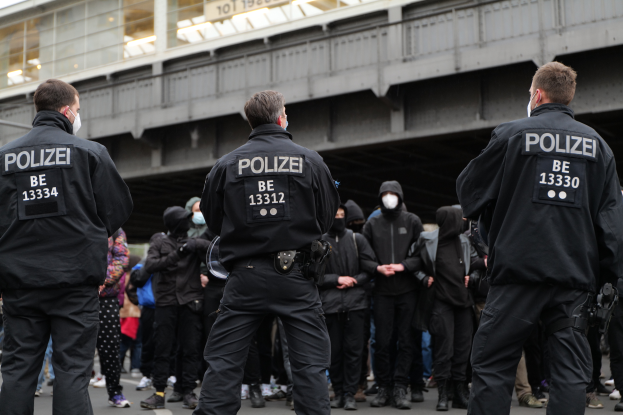 Eine Gruppe von Polizisten in Uniform steht vor einer Menge von Menschen in schwarzen Uniformen und Masken, mit einer Brücke und einem Gebäude im Hintergrund, während einer Demonstration in einer Stadt.