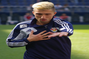 Ein junger Mann in einem blauen T-Shirt steht auf einem Fußballfeld und streckt die Arme aus, entschlossen als Flügelspieler mit einem unscharfen Hintergrund.