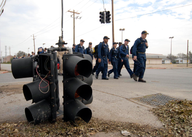Eine Gruppe uniformierter Polizisten geht eine Straße entlang neben einer Ampel, mit Strommasten, Gebäuden, Bäumen und einem klaren blauen Himmel im Hintergrund.
