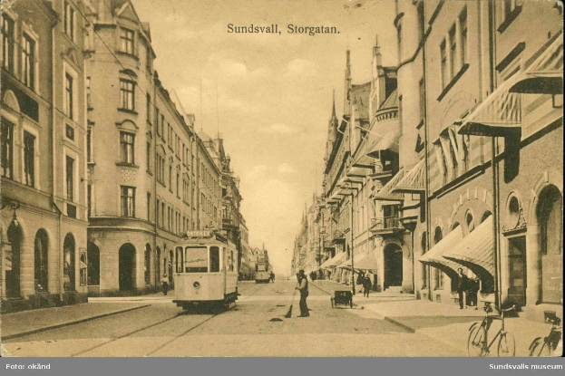 Ein altes Foto einer Stadtstraße mit einer Tram, Gebäuden, Fußgängern, Fahrrädern und einem bewölkten Himmel, mit Text oben und unten.