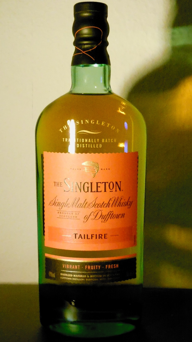 Eine tiefe Bernsteinflasche Singleton of Dufftown Tailfire Single Malt Scotch Whisky auf einem Tisch, mit einem schwarzen Etikett, das eine fette Schrift und ein Wappen mit einem Löwen und einer Burg zeigt.