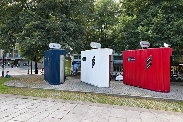 Drei mobile Toiletten auf einer grasbewachsenen Straßenecke mit Gebäuden und einem klaren blauen Himmel im Hintergrund.