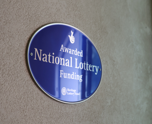 Blauer rechteckiger Schild an einem Gebäude mit weißem Hintergrund und schwarzer fetter Schrift, die "National Lottery Funding Awarded" lautet.