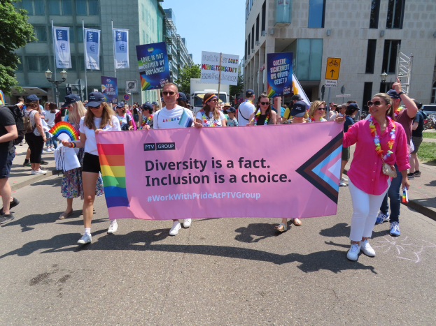 Eine bunte Gruppe von Menschen marschiert auf einer Straße, trägt ein Banner mit der Aufschrift "Diversität ist eine Tatsache, Inklusion ist eine Wahl", einige tragen Mützen und Schutzbrillen und andere zusätzliche Banner, vor einem Hintergrund von Gebäuden, Bäumen, Laternenpfählen und einem klaren blauen Himmel.
