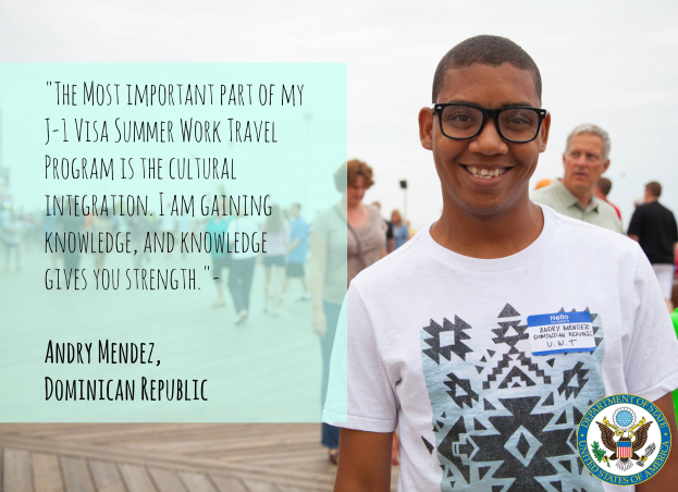 Eine Person in einem weißen T-Shirt lächelt beim Spaziergang mit einer Gruppe auf einem Holzbelag, mit einem Zitat über kulturelle Integration von Andry Mendez links, Gebäuden und einem klaren blauen Himmel im Hintergrund.