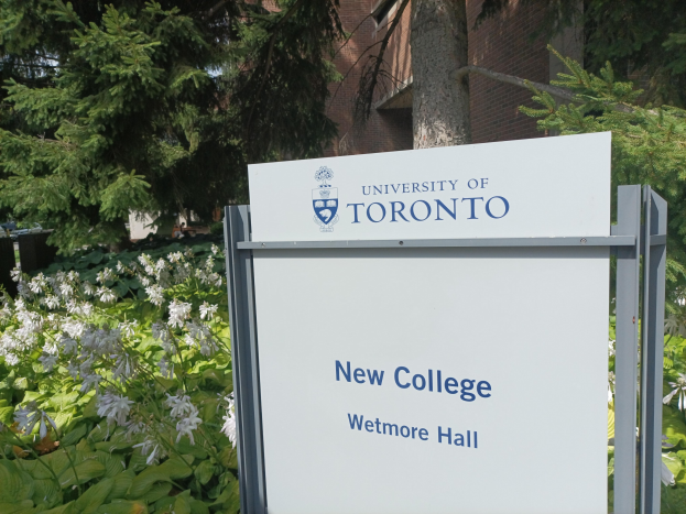 Außenansicht des neuen Colleges der Universität Toronto am Wetmore Hall, mit einem Schild vor dem Gebäude, umgeben von grünem Gras, bunten Blumen, hohen Bäumen und einem hellblauen Himmel.