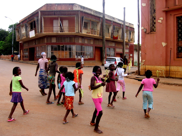 Eine Gruppe von Kindern geht eine Straße entlang, gesäumt von Gebäuden, Straßenlaternen, Straßenlaternen, Fahrzeugen, Bäumen und einem klaren blauen Himmel.