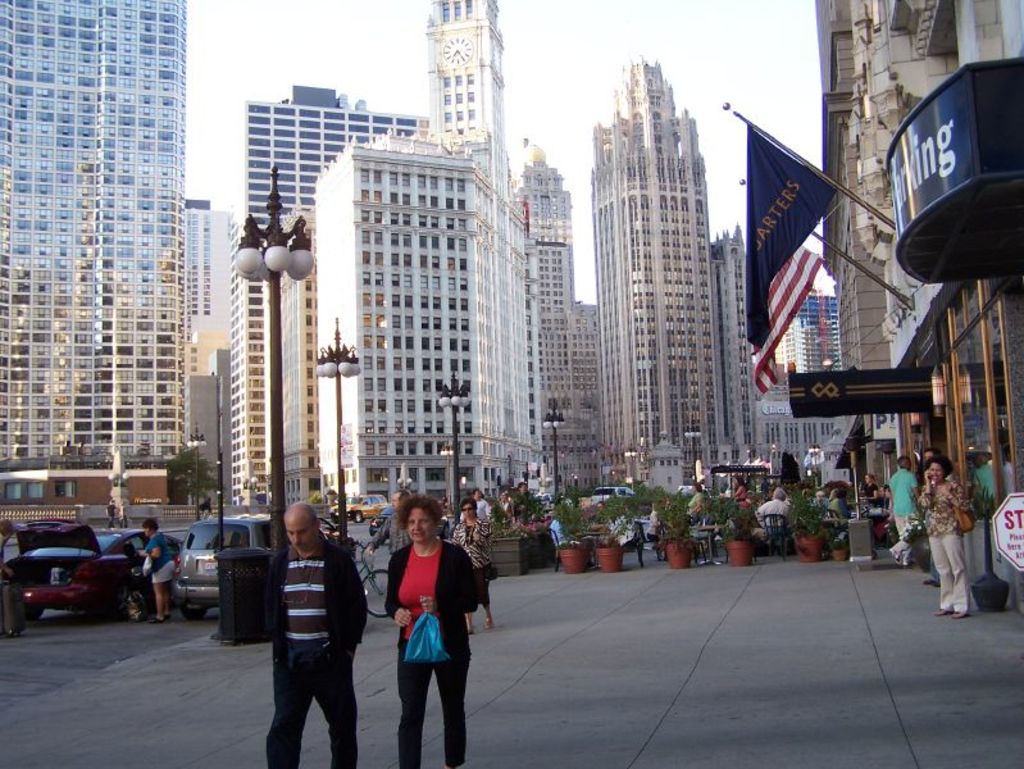 Eine belebte Straßenszene mit Menschen, Blumentöpfen, Autos, Straßenlaternen, Gebäuden im Hintergrund, Geschäften auf der rechten Seite und Fahnen.