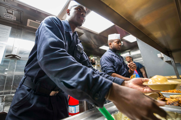 Eine Gruppe von Männern steht in einer Küche, einer hält einen Teller mit Essen, mit Tabletts mit Essen auf dem Tisch, einem Feuerlöscher und Papieren an der Wand im Hintergrund.