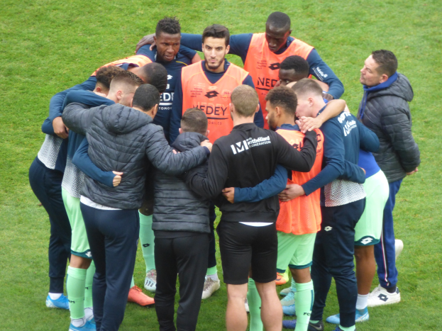 Eine Gruppe von Männern in Jacken, die sich auf einem Fussballfeld im Kreis versammelt haben.