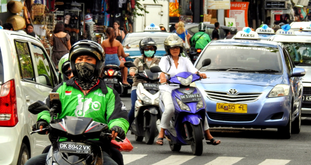 Eine Gruppe von Menschen auf Motorrädern fährt auf einer vielbefahrenen Straße mit Autos und anderen Fahrzeugen, einige Fahrer tragen Helme, im Hintergrund sind Gebäude, Bäume, Texttafeln und Geschäfte zu sehen.