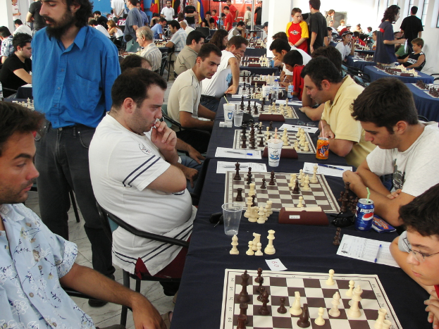 Eine Gruppe von Menschen spielt Schach mit einem Mann in einem blauen Hemd auf der linken Seite, ein Fenster in der oberen rechten Ecke und ein Tisch mit einem Glas, einem Schachbrett, Schachfiguren, einer Coke, Papier, Stift und einer Tischdecke.