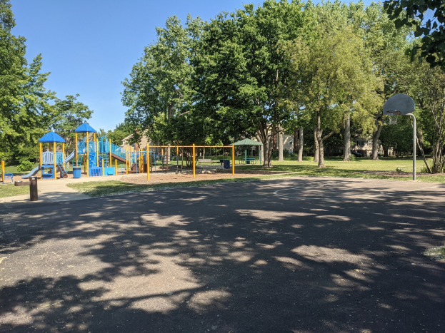 Ein Park mit einem Spielplatz in der Mitte, umgeben von Bäumen unter einem klaren blauen Himmel, eine Straße unten und ein Basketballkorb rechts.