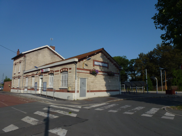 Ein Bahnhof mit einer Fußgängerüberquerung davor, umgeben von Gebäuden, Straßenlaternen, Strommasten, Stromkabeln, Kraftfahrzeugen, Bäumen und einem klaren blauen Himmel.