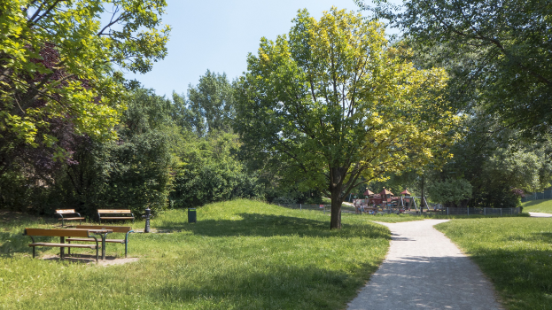 Ein Parkweg mit Bänken gesäumt, von grünem Gras und Bäumen umgeben, mit Hütten und einem klaren blauen Himmel im Hintergrund.