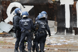 Polizeibeamte in schwarzen Uniformen und blauen Helmen stehen vor einem mit Graffiti bedeckten Gebäude während einer Ausschreitung, mit verstreuten Papieren und Gegenständen auf dem Boden.