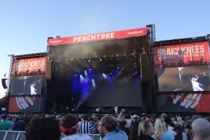 Eine große Menschenmenge vor einer hell erleuchteten Bühne beim Shaky Knees Music Festival, auf der Musiker unter Bannern spielen, mit einem Geländer im Vordergrund und einem klaren blauen Himmel dahinter.