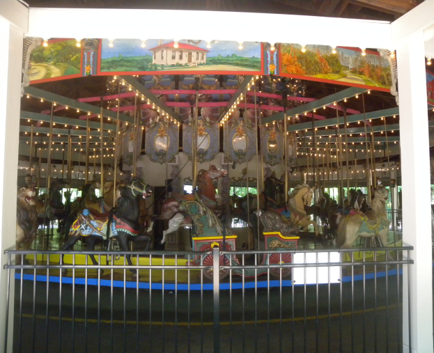 Ein hell erleuchtetes Karussell mit Pferden, umgeben von einem Geländer und Säulen, in einem Gebäude, das eine festliche Atmosphäre schafft.