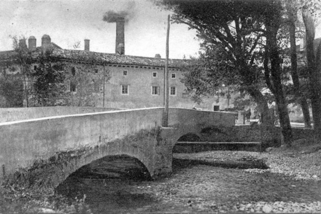 Stadtansicht mit Gebäuden, Bäumen und einer Brücke im Vordergrund, Rauch aus einem Schornstein, unter einem Himmel mit Wasser am Boden.