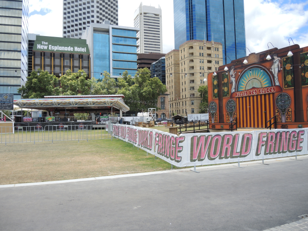 Ein Karnevalsritt wird in einer Stadtstraße aufgebaut, umgeben von hohen Gebäuden, Bäumen und einem bewölkten Himmel, mit einer Straße, einem Geländer und einer Banner mit Text im Vordergrund.