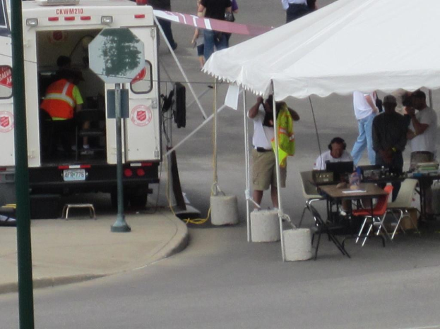 Ein Rettungswagen steht links neben einer Straße, während auf der rechten Seite ein Zelt eine Gruppe von Menschen schützt, mit einem Tisch und einem Stuhl in der Nähe.