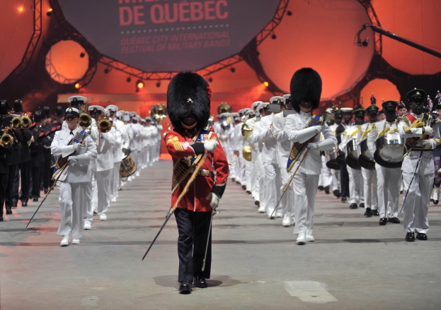 Eine Gruppe uniformierter Personen marschiert auf einer Straße, einige spielen Musikinstrumente, mit einem beleuchteten Schild im Hintergrund, das die Eröffnungszeremonie des Montreal International Festival of Military Bands anzeigt.