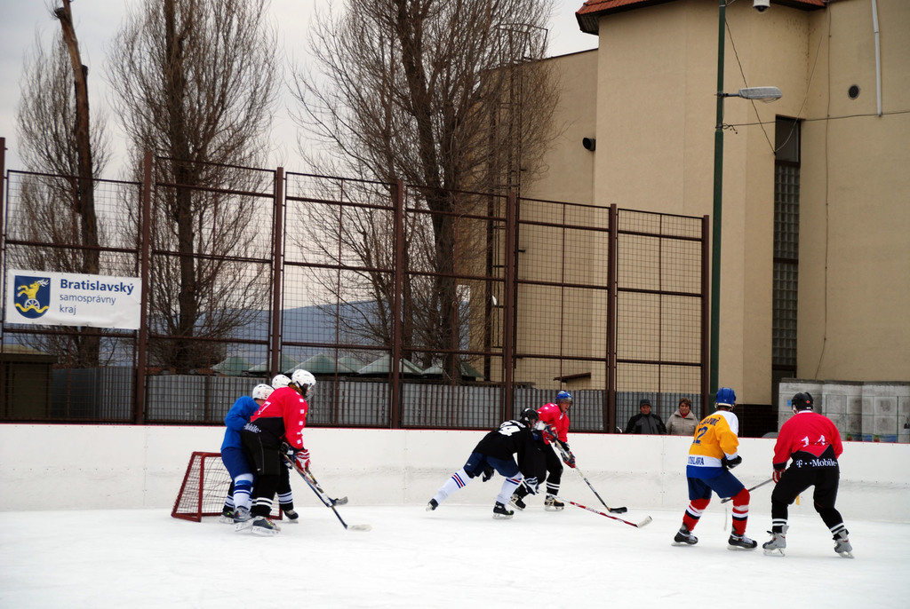 Menschen spielen Eishockey auf einer Eisfläche mit einem Hintergrund aus Himmel, Bäumen, Gebäuden, einer Straßenlaterne, einem Namensschild und Zäunen.