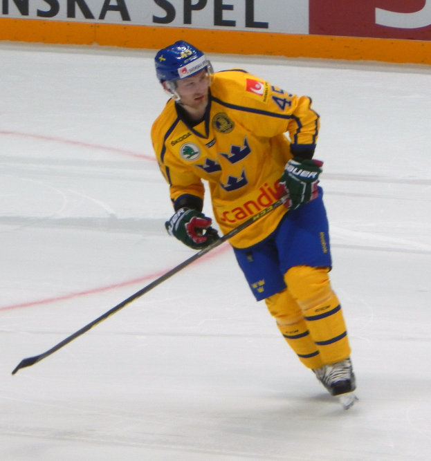 Eishockeyspieler in Bewegung auf dem Eis mit Helm, Handschuhen und Schlittschuhen, der einen Hockeystock hält, mit einem Hintergrundboard mit Text.