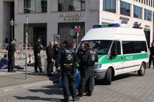Eine Gruppe von Polizisten steht vor einem Starbucks-Café, mit einem Van auf der rechten Seite und einigen Menschen auf der linken Seite, sowie einem Gebäude mit Fenstern, einem Schild, einem Laternenpfahl und einem Zaun im Hintergrund.