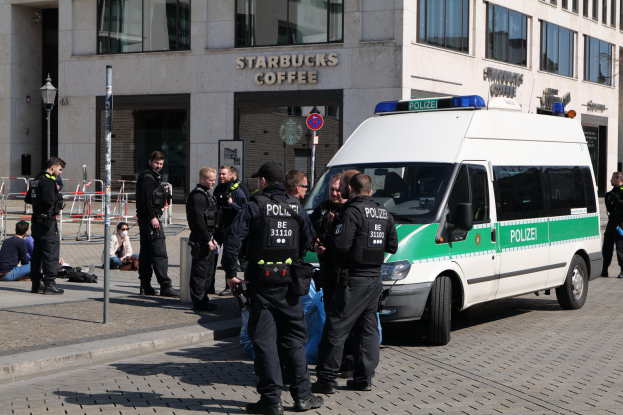 Eine Gruppe von Polizisten steht vor einem Starbucks-Café, mit einem Van auf der rechten Seite und einigen Menschen auf der linken Seite, sowie einem Gebäude mit Fenstern, einem Schild, einem Laternenpfahl und einem Zaun im Hintergrund.