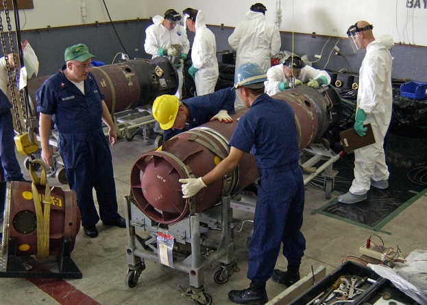 Eine Gruppe von Männern in Helmen und Handschuhen bei der Entschärfung einer großen Bombe in einer Fabrik mit Maschinen und einer Wandtafel im Hintergrund.