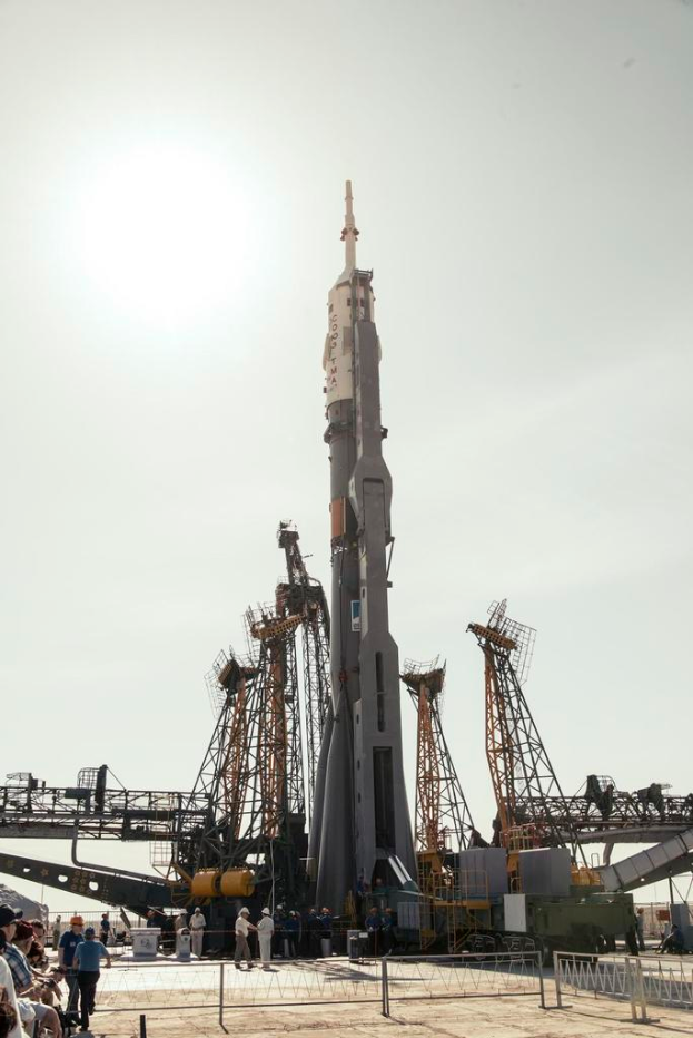 Ein Metallstabzaun mit ein paar Menschen links daneben, dahinter stehen Ingenieure vor einer Raketenstartrampe mit einer Rakete und diverser Ausrüstung.