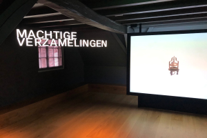 Installationsansicht der Ausstellung 'Machtige Sammlung' im Museum of Modern Art in Berlin, mit einem großen Bildschirm auf der rechten Seite und Text an der Wand dahinter sowie einem Fenster auf der linken Seite.