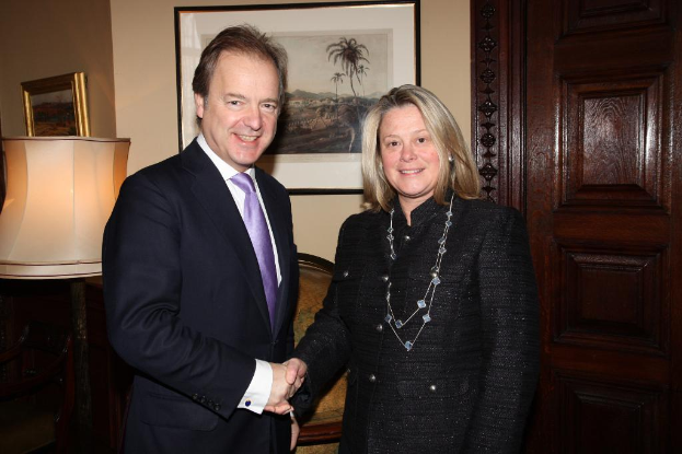 Eine Frau und ein Mann in einem Anzug l├Ącheln, geben sich die Hand und schauen sich an, mit einer Wand, Fotorahmen, einer Lampe, einer T├╝r und ein paar Gegenst├Ąnden im Hintergrund.