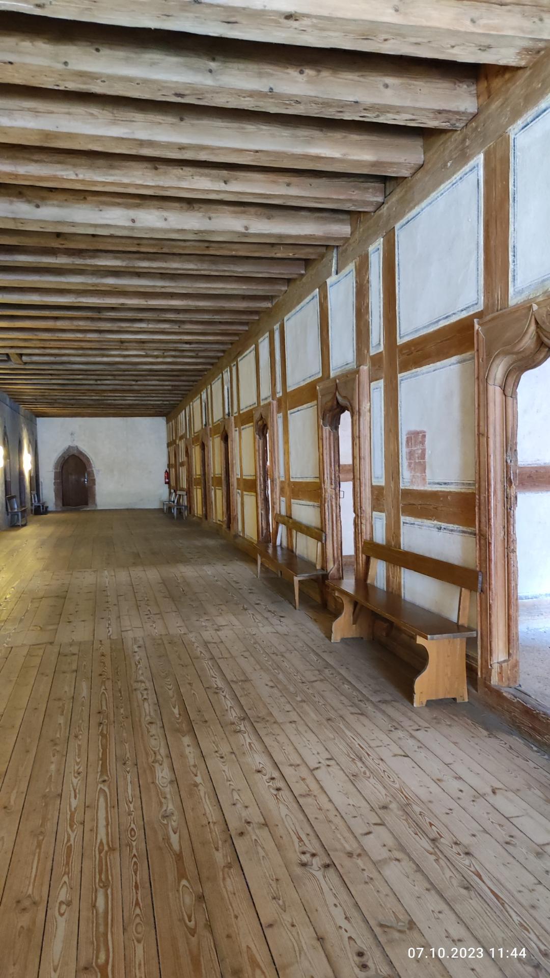 Ein geräumiger Raum mit Holzboden, weißen Wänden, Bänken, Fenstern, Lampen und einer hohen Decke, wahrscheinlich der Hauptsaal eines Schlosses.