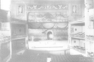 Schwarz-weiß-Fotografie des Auditoriums des Theatre Royal London, das eine Bühne mit Stühlen, Geländern, skulptierten Wänden und einem Vorhang zeigt, mit Text am unteren Rand.