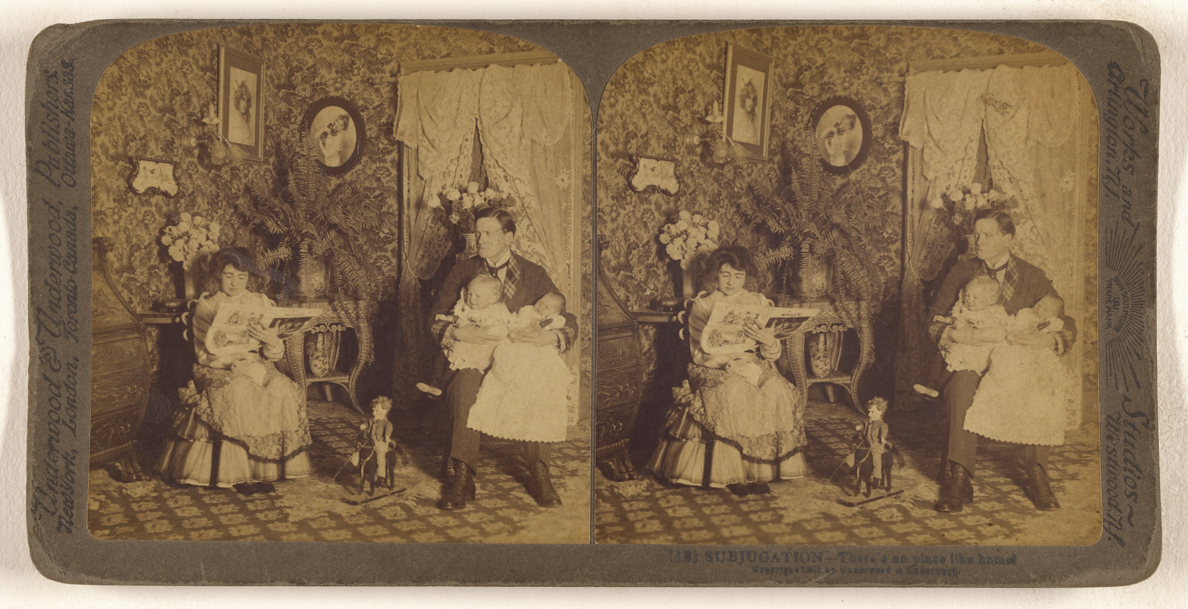 Altes Schwarz-Weiß-Foto einer Familiengruppe mit einem sitzenden Mann und einer Frau, die Babys halten, umgeben von Blumentöpfen, Vorhängen und gerahmten Bildern an der Wand.