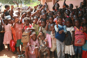 Eine Gruppe von Kindern steht mit erhobenen Händen, mit einem Gebäude auf der rechten Seite und einem Fahrzeug unter Bäumen im Hintergrund.