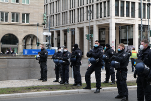 Eine Gruppe von Polizisten in schwarzen Uniformen und Masken steht vor einem Glasfenstergebäude mit Säulen in Berlin, Deutschland, einige halten Helme, mit Laternen, Verkehrsampeln, Schildern mit Text, einer Statue, anderen Menschen und grasbewachsenem Boden im Hintergrund.
