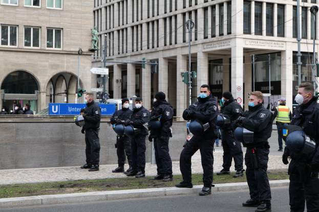 Eine Gruppe von Polizisten in schwarzen Uniformen und Masken steht vor einem Glasfenstergebäude mit Säulen in Berlin, Deutschland, einige halten Helme, mit Laternen, Verkehrsampeln, Schildern mit Text, einer Statue, anderen Menschen und grasbewachsenem Boden im Hintergrund.