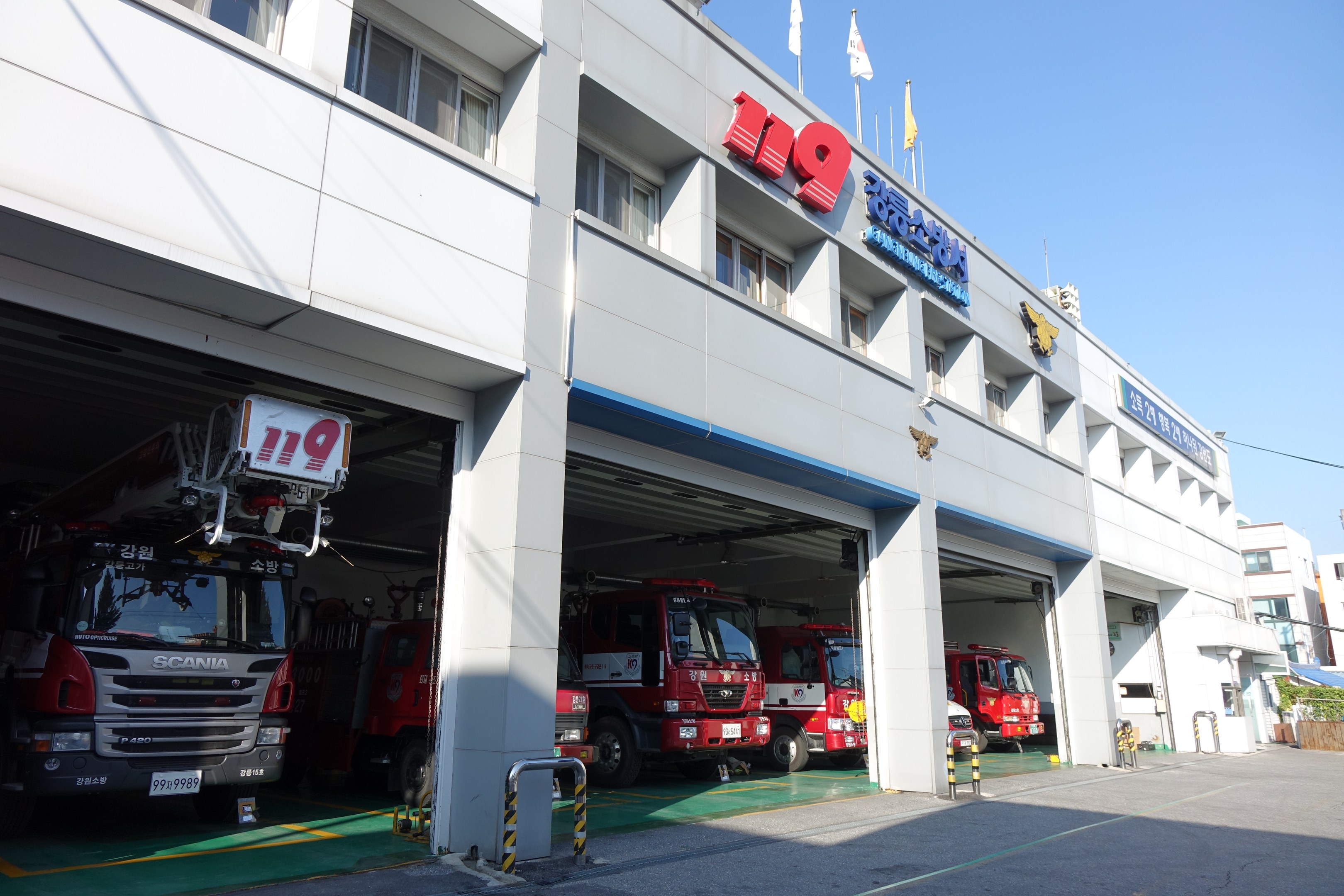 Feuerwache mit mehreren Feuerwehrautos vor dem Gebäude geparkt, mit Fenstern, Säulen, Namensschildern und Flaggen im Vordergrund auf Stangen, vor anderen Gebäuden unter einem klaren blauen Himmel.