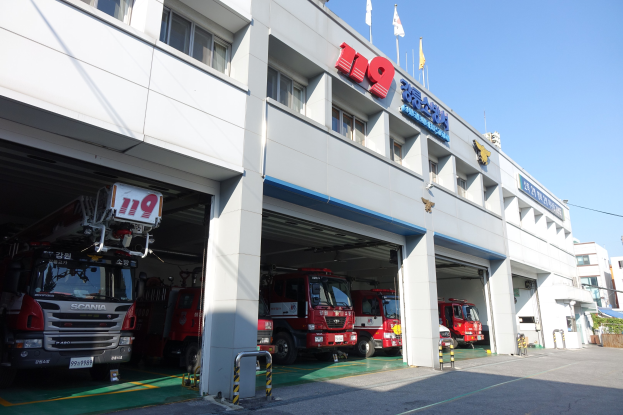 Feuerwache mit mehreren Feuerwehrautos vor dem Gebäude geparkt, mit Fenstern, Säulen, Namensschildern und Flaggen im Vordergrund auf Stangen, vor anderen Gebäuden unter einem klaren blauen Himmel.