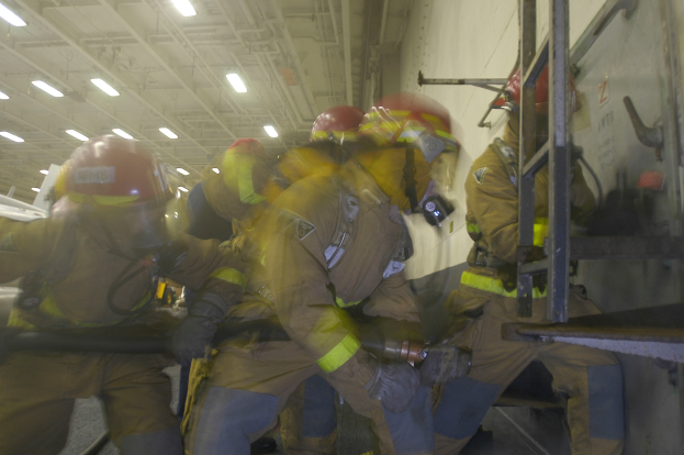 Feuerwehrleute in Helmen und Schutzausrüstung arbeiten an einem Flugzeug, halten verschiedene Gegenstände und haben eine Leiter auf der rechten Seite, unter Deckenlampen.