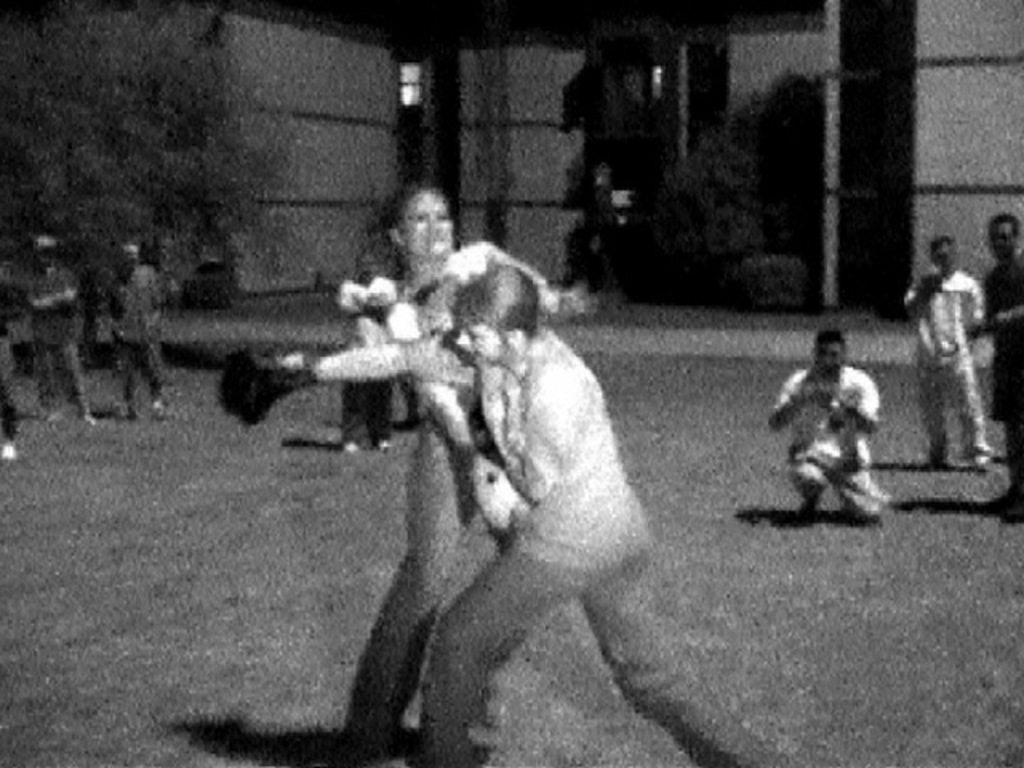 Zwei Personen boxen in der Mitte eines Schwarz-Weiß-Bildes, im Hintergrund sind ein Gebäude und einige Zuschauer zu sehen.