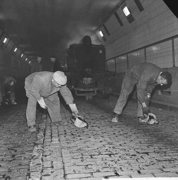 Schwarzes und weißes Bild von Männern, die auf einem Ziegelboden in einem Tunnel arbeiten, mit einem Zug im Hintergrund und Deckenbeleuchtung.