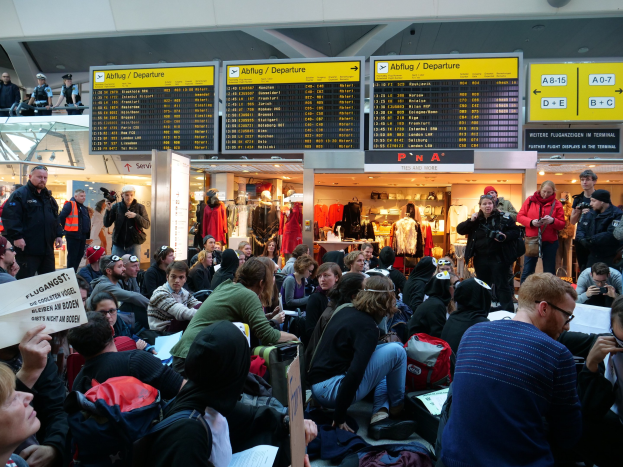 Eine große Gruppe von Menschen an einem Flughafen, einige mit Taschen und Papieren sitzend, andere stehend, mit Texttafeln, Schaufensterpuppen in Kleidern und Deckenlampen im Hintergrund, was auf eine Demonstration hinweist.